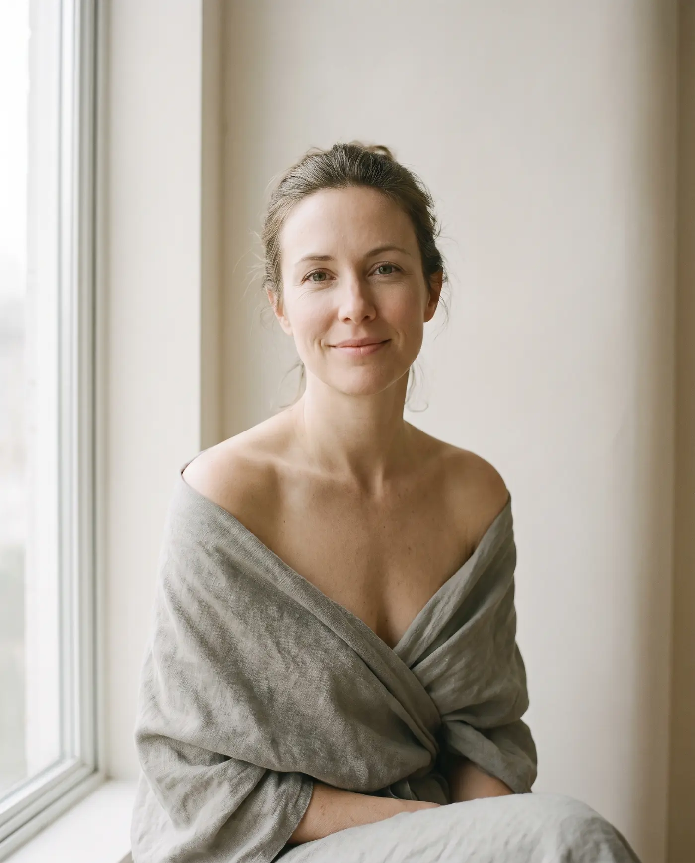 A woman seated in soft window light, resting her gaze on the camera